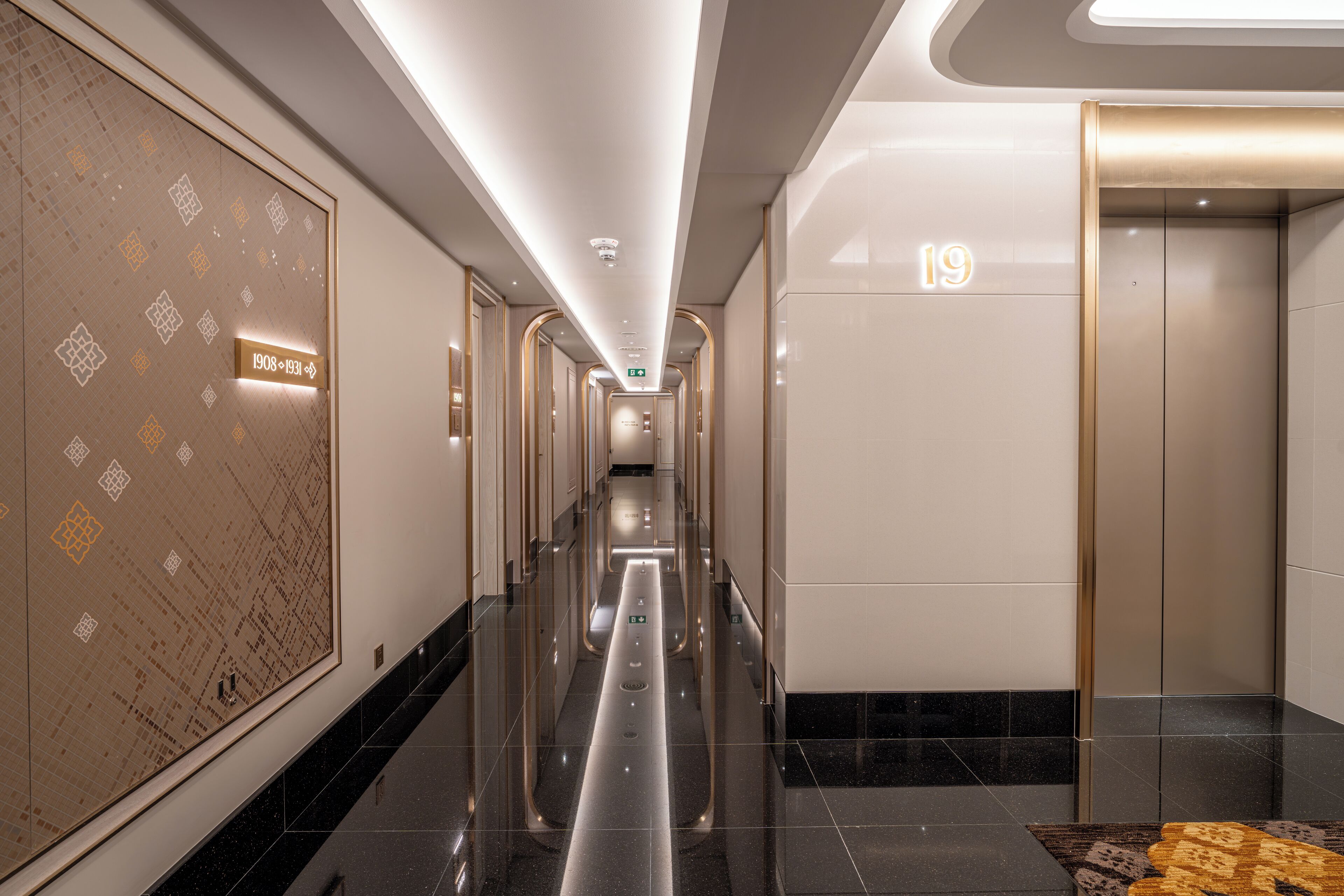 A modern hotel corridor with a polished floor, a patterned wall, and an elevator marked with the number 19.