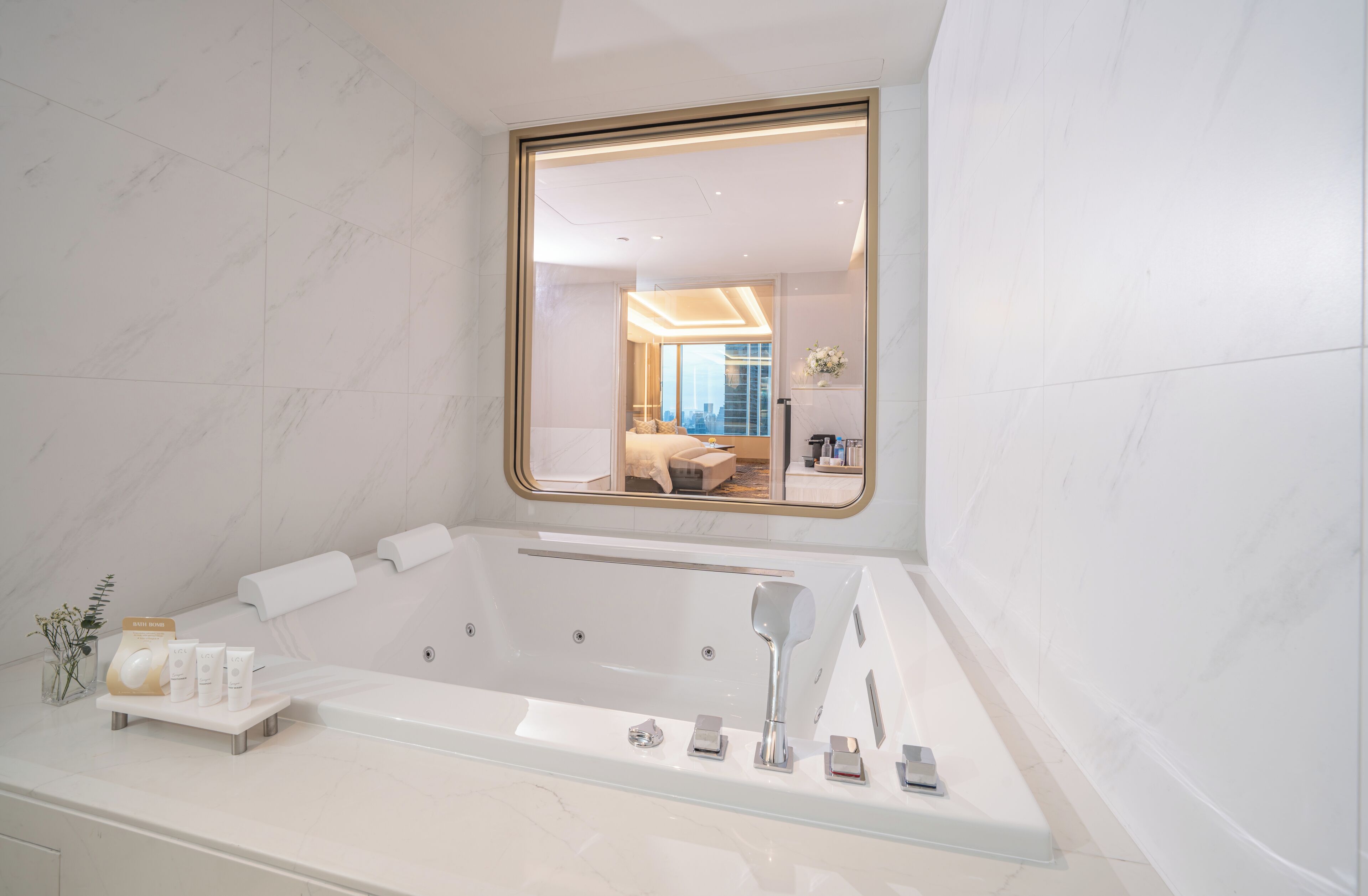 A modern bathroom with a large bathtub, a vanity with toiletries, and a mirror reflecting a bedroom with a bed and a city view.
