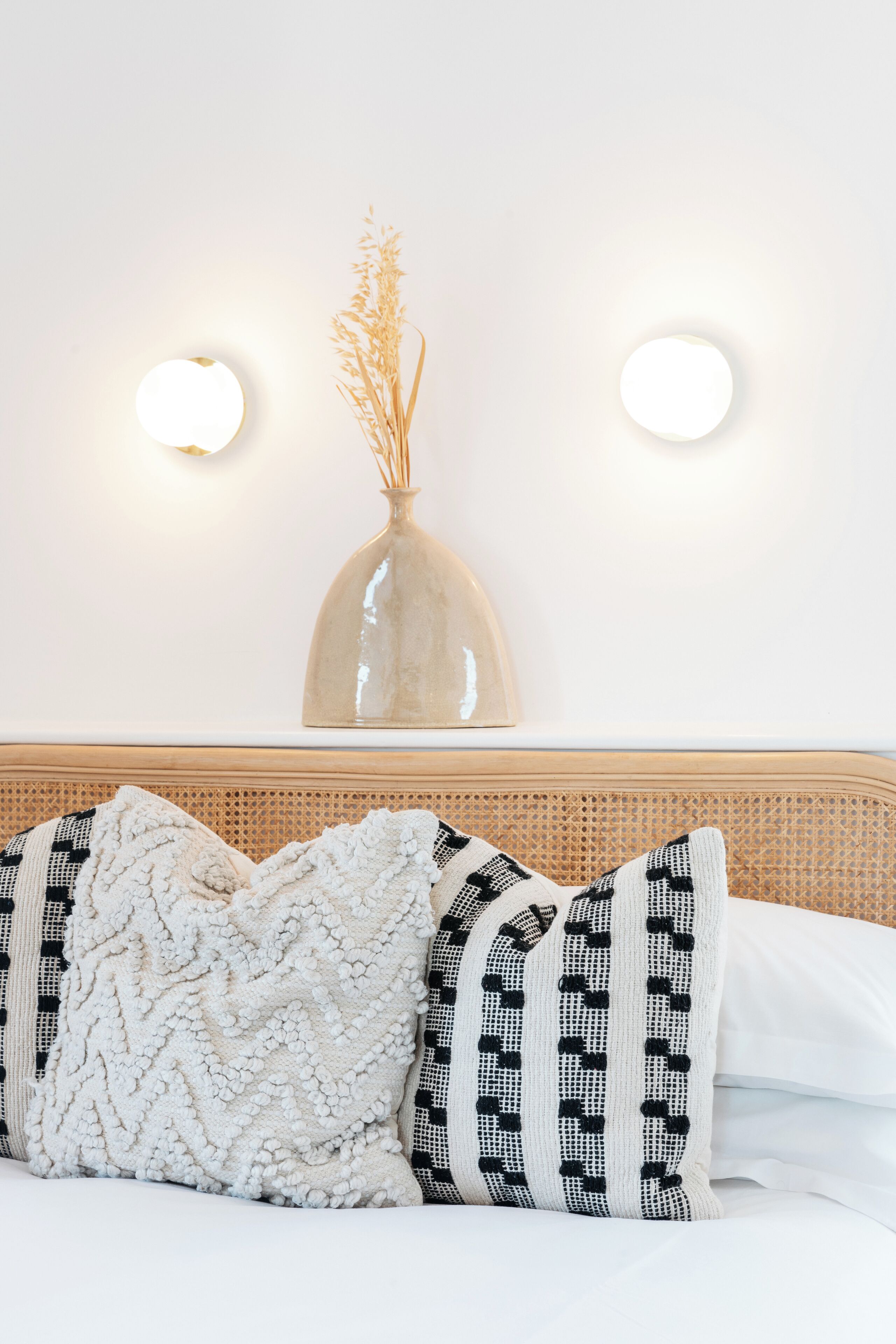 A bed with white and black patterned pillows, a woven headboard, and a vase with dried plants on a wall.