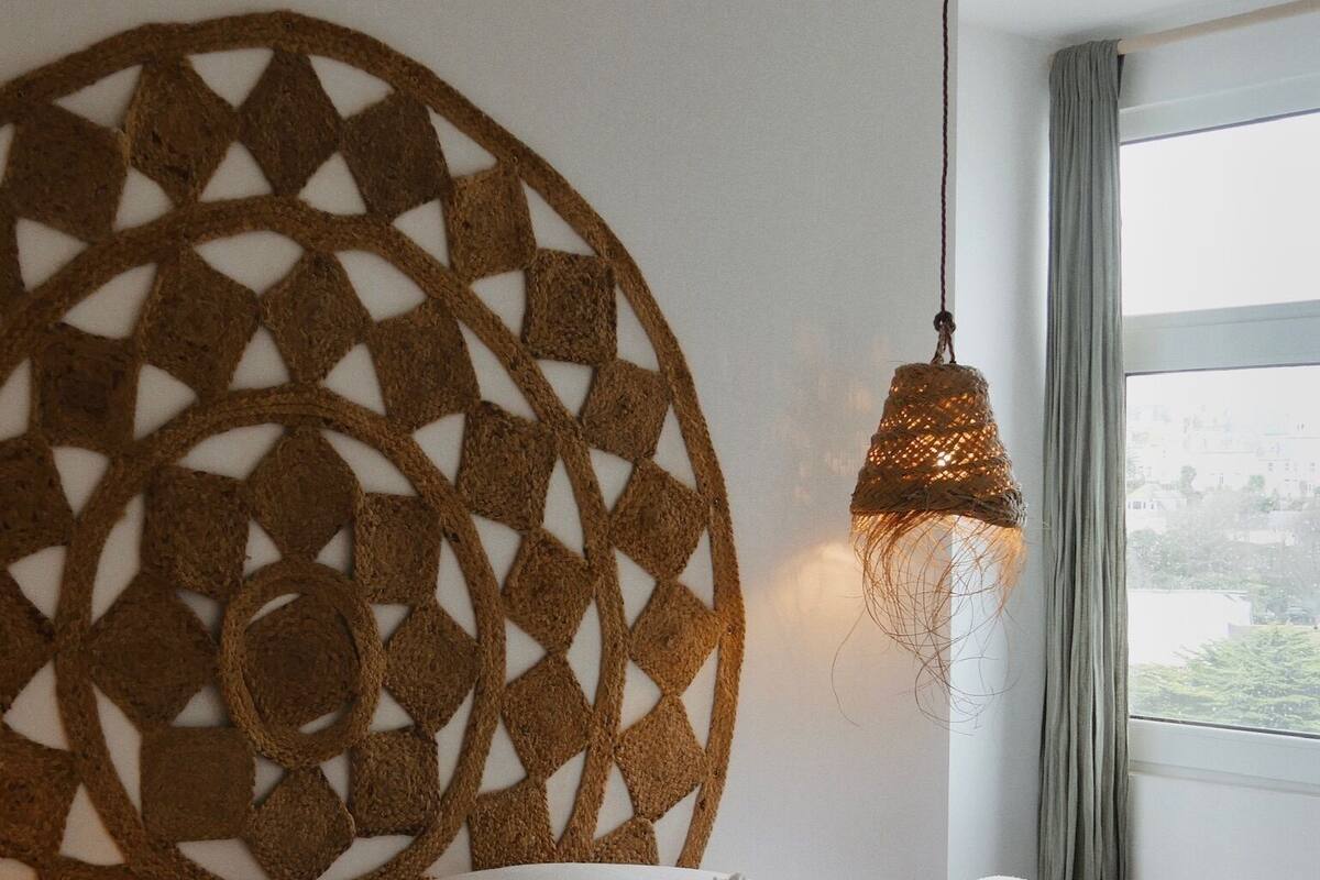 A bedroom with a large, circular woven headboard, a hanging light fixture, and a view of greenery through the window.