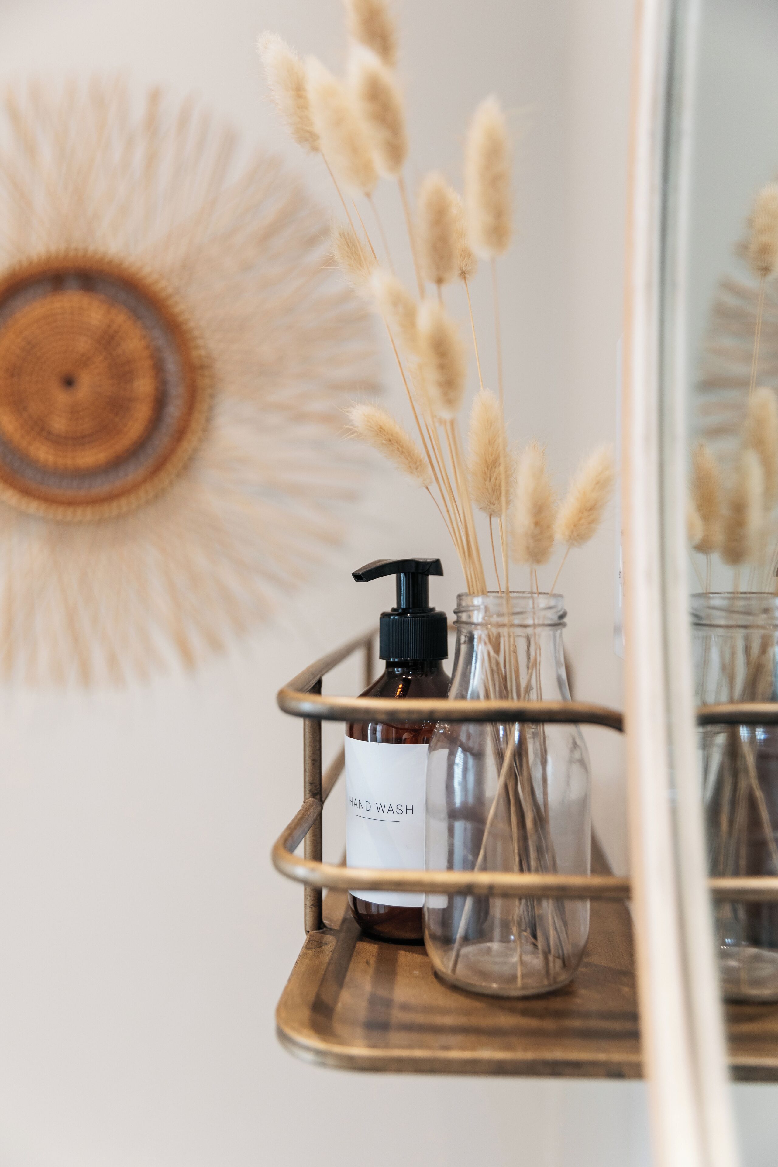 A bathroom vanity with a mirror, a wooden tray, a glass jar with pampas grass, and a bottle with a pump.