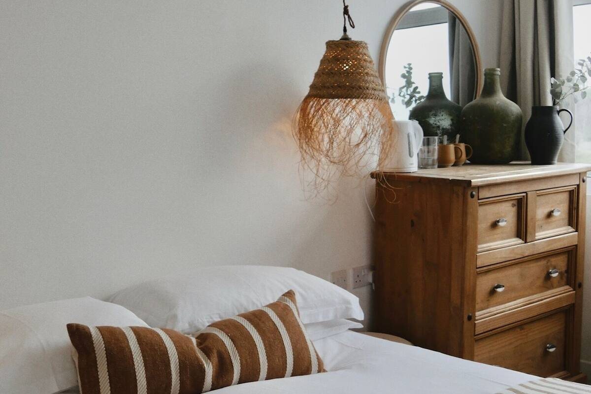 A neatly made bed with striped bedding, a hanging woven light fixture, a wooden dresser with drawers, and a mirror on the wall.
