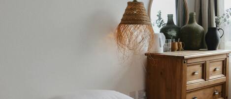 Una cama bien tendida con ropa de cama a rayas, una lámpara colgante de fibra, un aparador de madera con cajones y un espejo en la pared.
