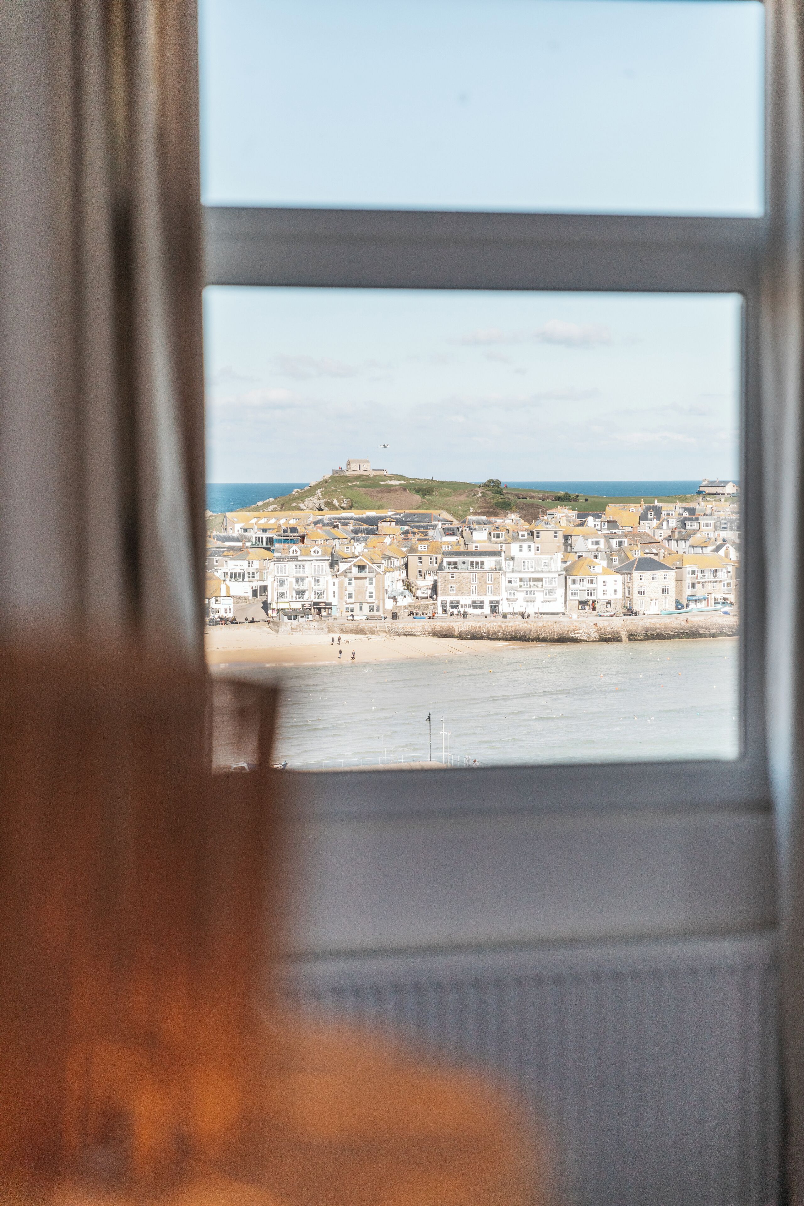 A coastal town view through a window, featuring buildings, a beach, and a hill with a structure on top.