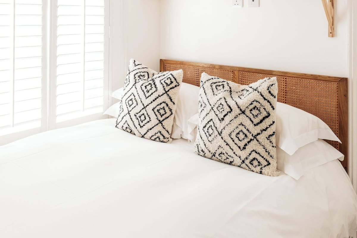 A bedroom with a bed, white bedding, patterned pillows, a wooden headboard, a window with blinds, and a shelf with decorative items.