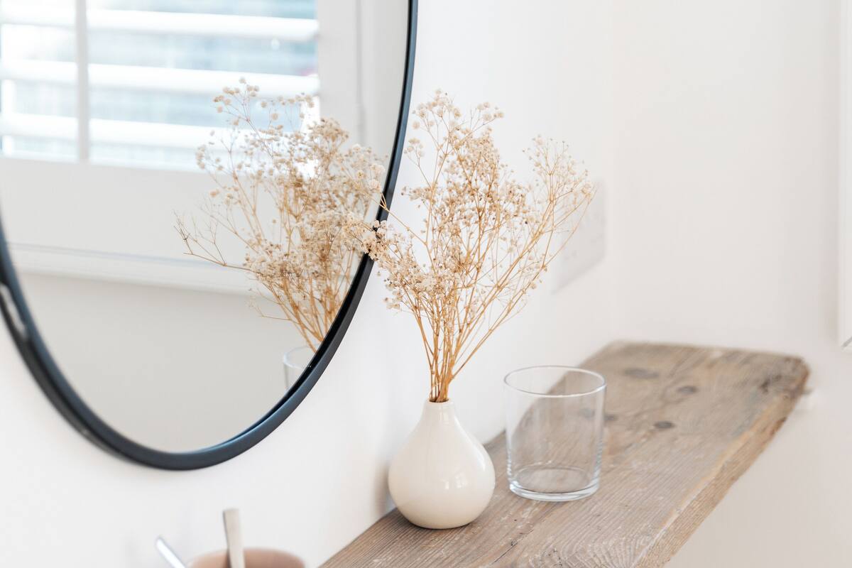 A wooden shelf with a white vase containing dried flowers, a clear glass, and a pink mug with a spoon.