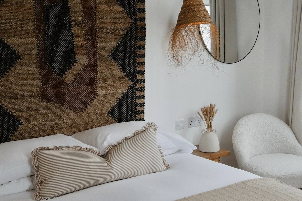 A neatly made bed with white linens and striped pillows, a large woven headboard, a round mirror, and a hanging light fixture.
