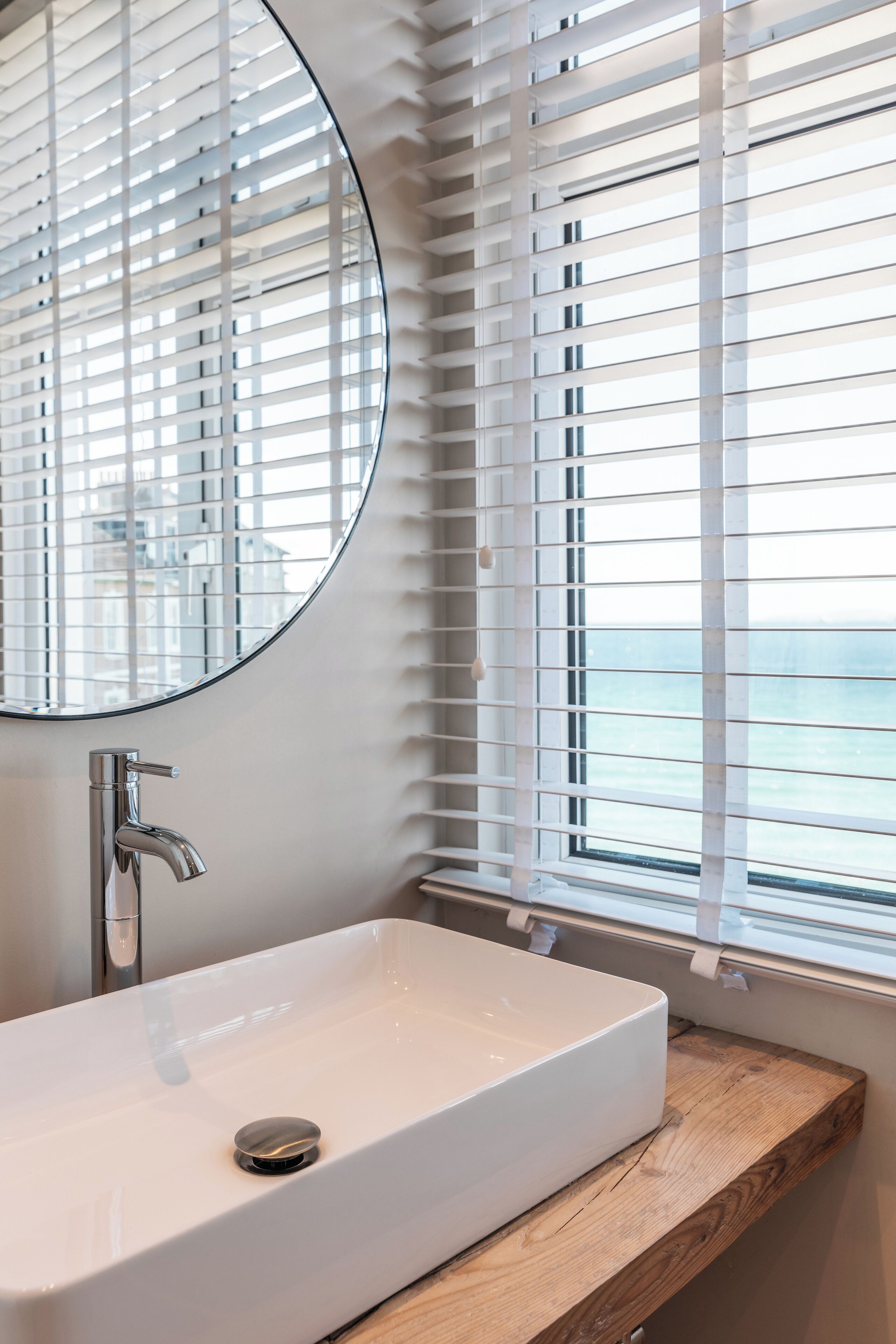 A bathroom with a white sink, a round mirror, and a window with blinds.