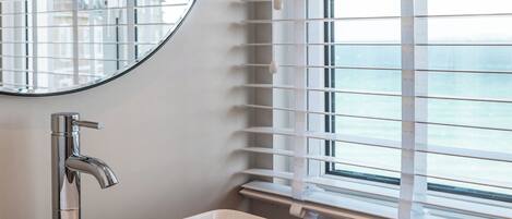 A bathroom with a white sink, a round mirror, and a window with blinds.