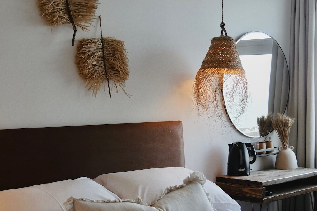 A neatly made bed with white linens and a dark green throw blanket. There are two decorative wall hangings made of straw-like material. A round mirror is visible on a wooden nightstand beside the bed, which also holds a black coffee maker and a white vase with dried plants.
