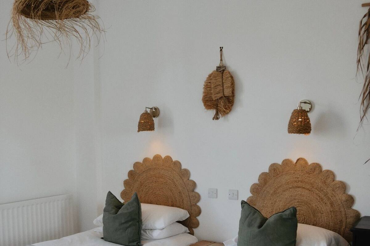 A room with two beds, each with a scalloped headboard, white bedding, and green pillows. There are wall-mounted light fixtures and a woven hanging light fixture.