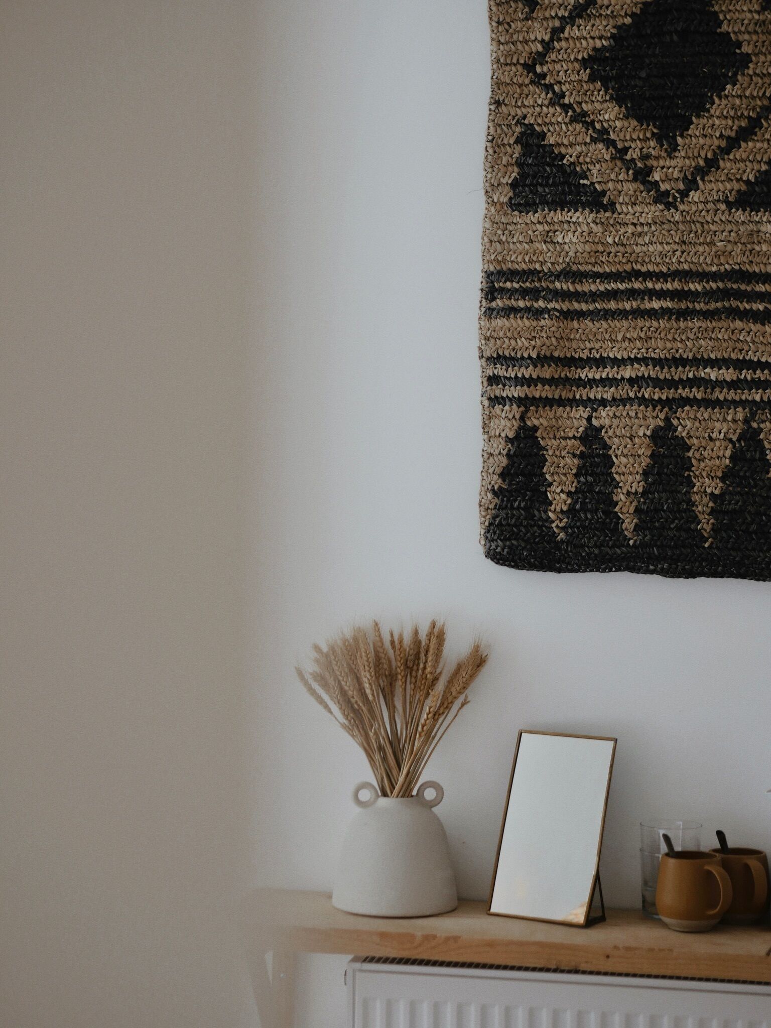 A wall with a patterned tapestry, a white vase with dried plants, a framed picture, and two mugs on a wooden shelf.