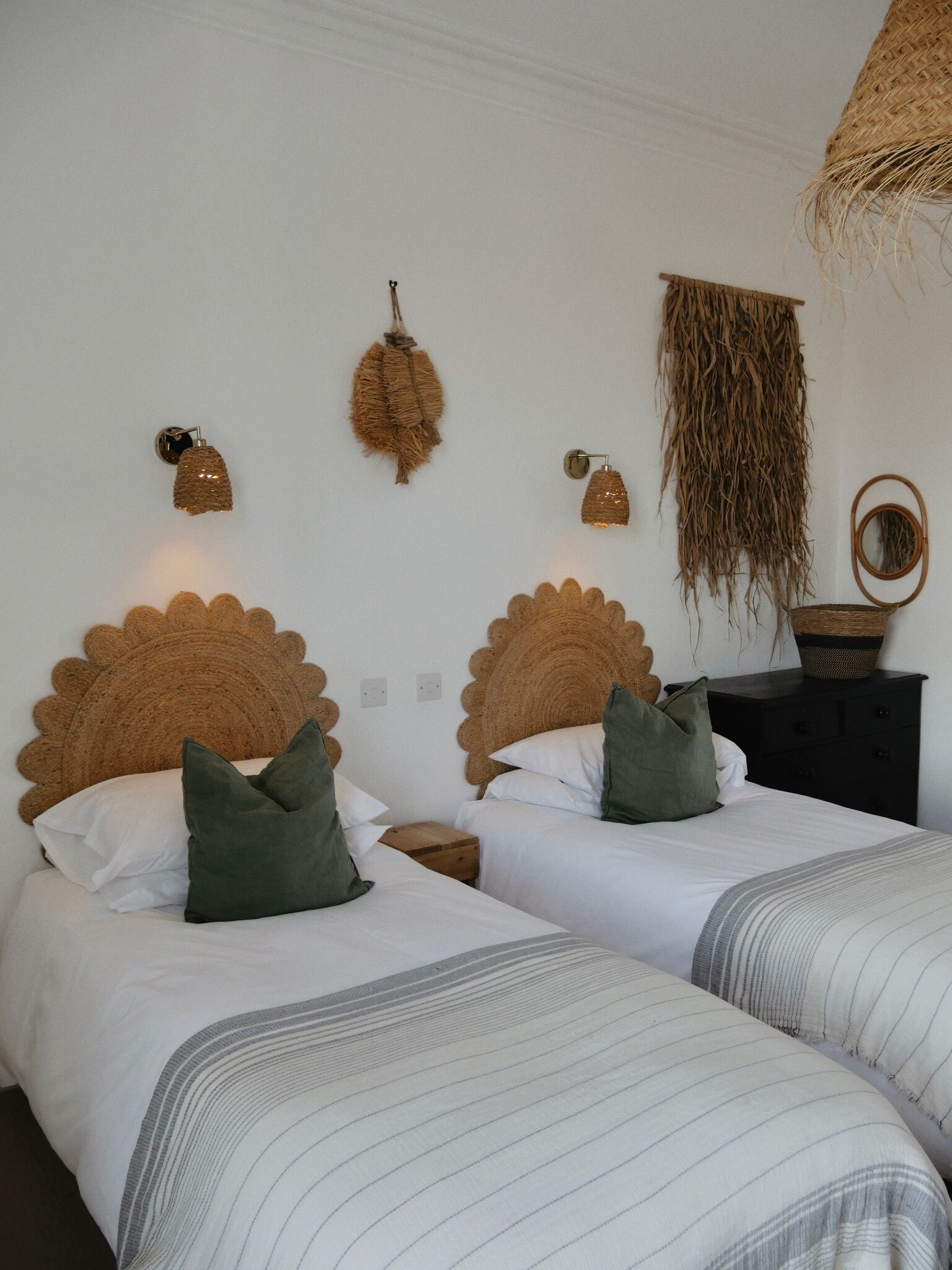 A bedroom with two beds, each with a unique headboard design. There are wall-mounted lights and a woven basket on a nightstand.