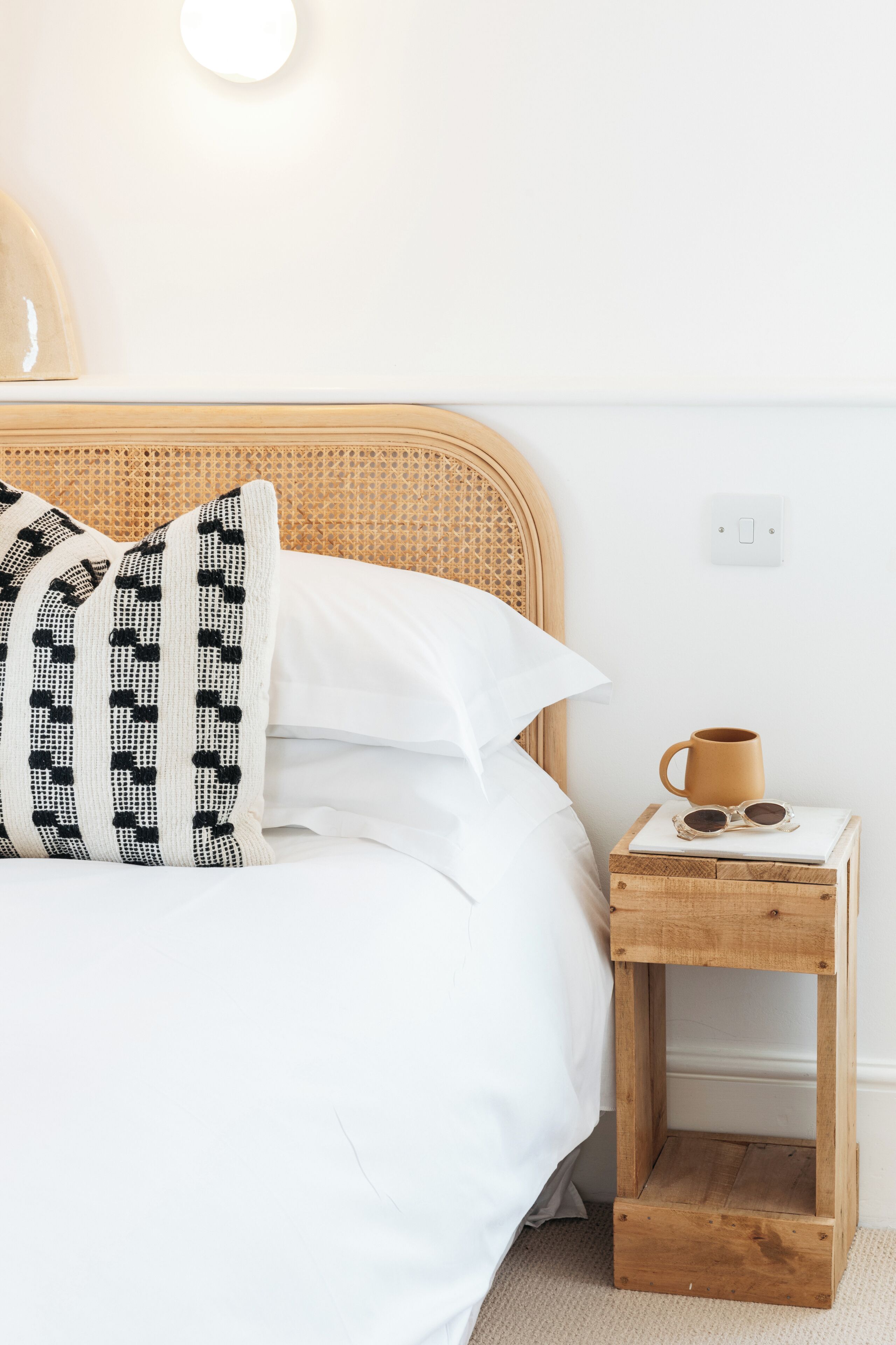 A neatly made bed with white linens and a black and white patterned pillow. A wooden nightstand with a mug and sunglasses is beside the bed.