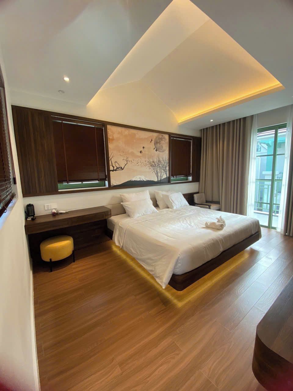 A modern hotel room with a large bed, wooden flooring, a painting on the wall, and a window with curtains.