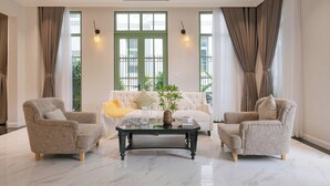 Un salon spacieux avec un canapé blanc, deux fauteuils, une table basse en verre et de grandes fenêtres avec des rideaux.
