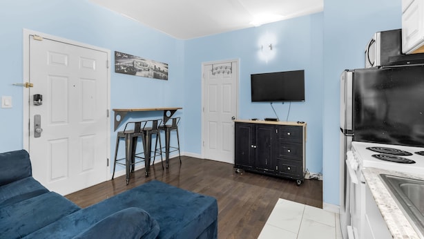 A living room with a blue sofa, a dining area with a table and chairs, a kitchen with a stove and refrigerator, and a TV mounted on the wall.