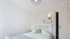 Une chambre avec un lit blanc, un plaid vert, une tête de lit en bois, une table de chevet avec une lampe et un miroir au mur.