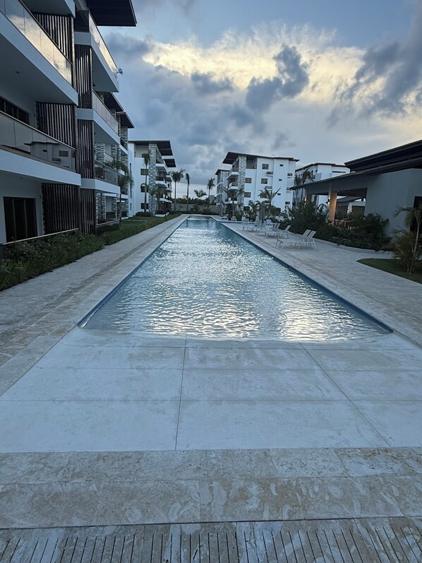 A modern residential building with a central swimming pool, surrounded by greenery and other buildings.