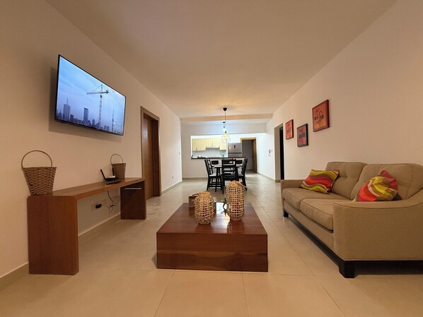 A modern living room with a flat-screen TV, a wooden coffee table, a sofa, and a dining area in the background.