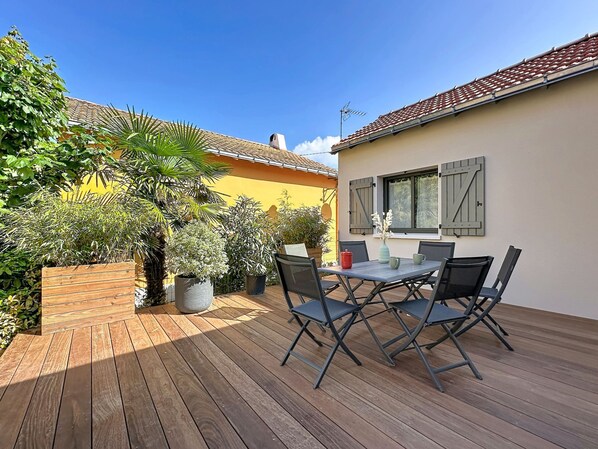 Une terrasse en bois avec une table et des chaises, entourée de plantes en pot et d’un mur jaune.