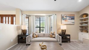 A living room with a beige sofa, two side tables with lamps, a coffee table with decorative bowls, a painting on the wall, and a window with curtains. - Glendale Paradise | Pool | Pickleball | Putt-Putt (Glendale)