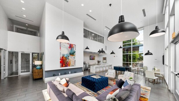 A spacious living area with a large sofa, a blue ottoman, and a colorful rug. There are multiple pendant lights and a large abstract painting on the wall. - Stylish Resort Oasis: 5* Location, Pool, Gym, Pkg! (San Antonio)