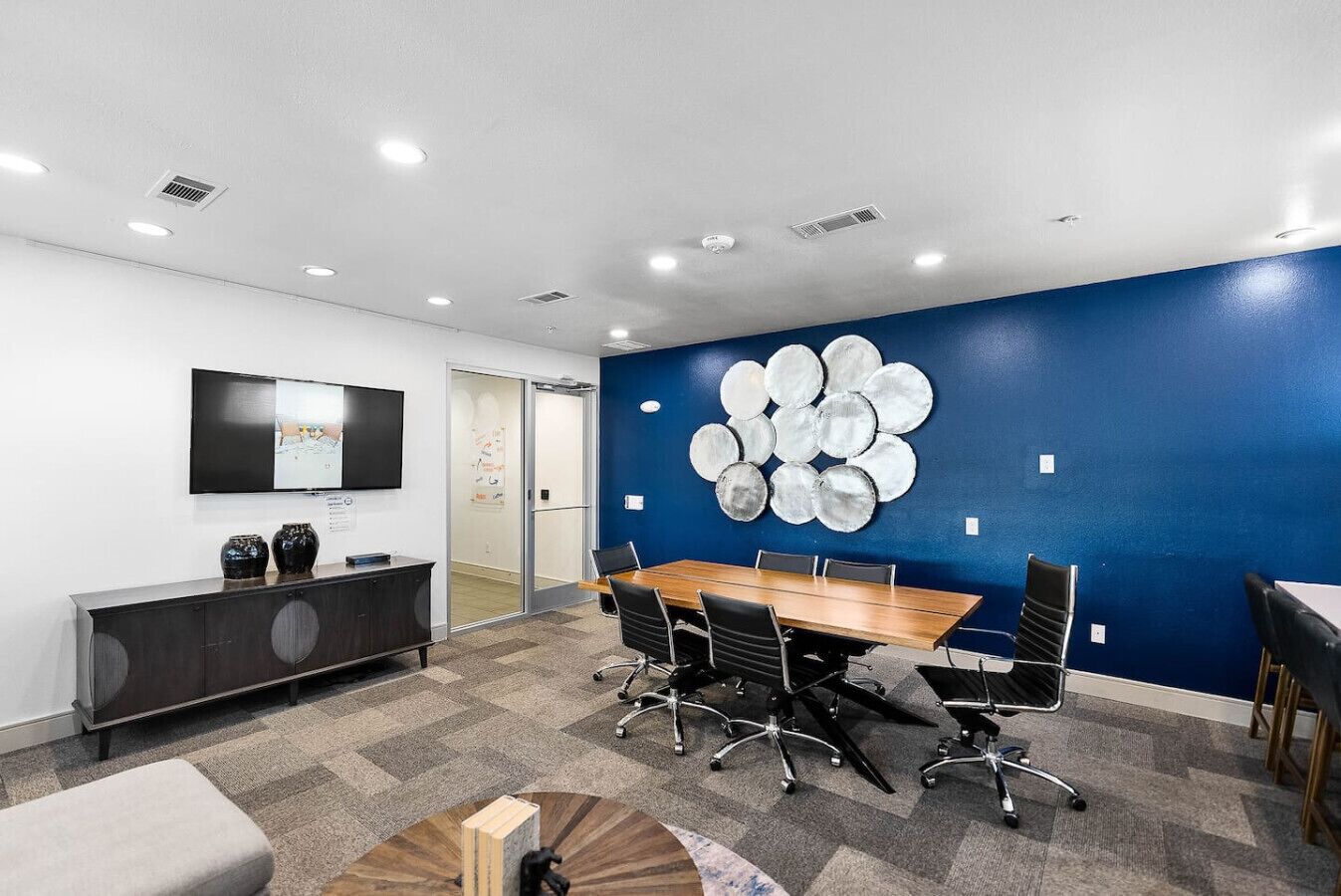 Una sala de conferencias con una gran mesa, sillas, un televisor en la pared y una pared azul decorada con un diseño circular.