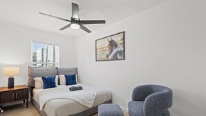 Une chambre à coucher comprenant un lit, une chaise, une table de chevet, une lampe, un ventilateur de plafond, une fenêtre et un tableau accroché au mur.