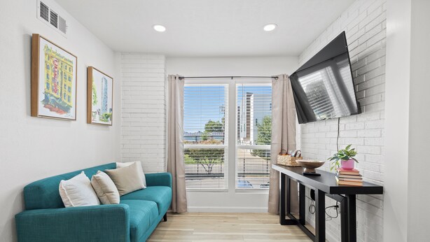 Un salon moderne avec un canapé de couleur turquoise, un téléviseur à écran plat fixé sur un mur en briques blanches et une vue sur la ville grâce à de grandes fenêtres.