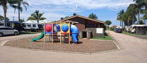 A playground with a slide and climbing structures, surrounded by palm trees and RVs parked nearby.