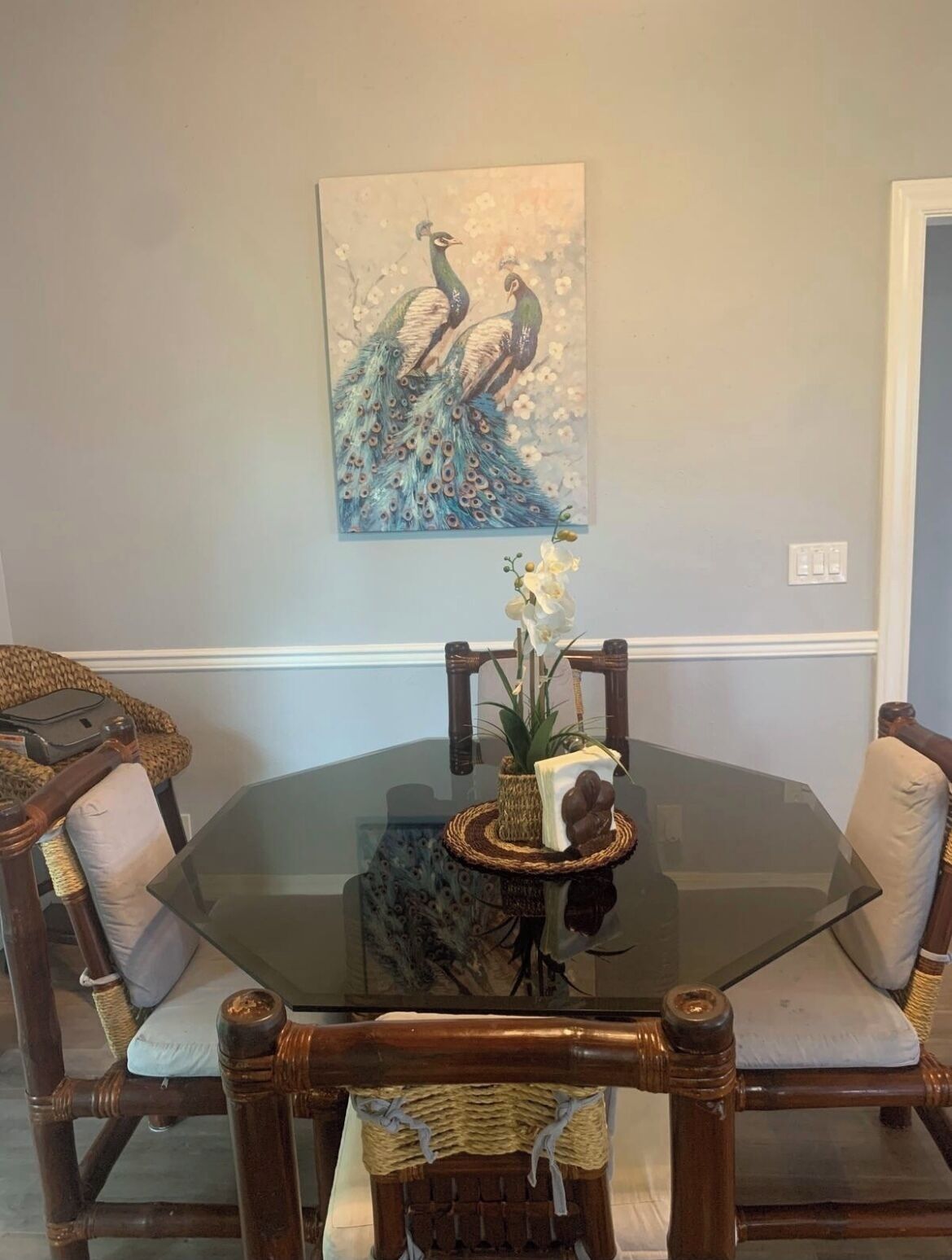 A dining area with a glass table surrounded by wicker chairs and a peacock painting on the wall.