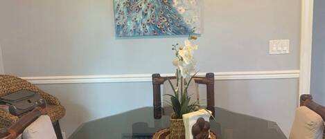 A dining area with a glass table surrounded by wicker chairs and a peacock painting on the wall.