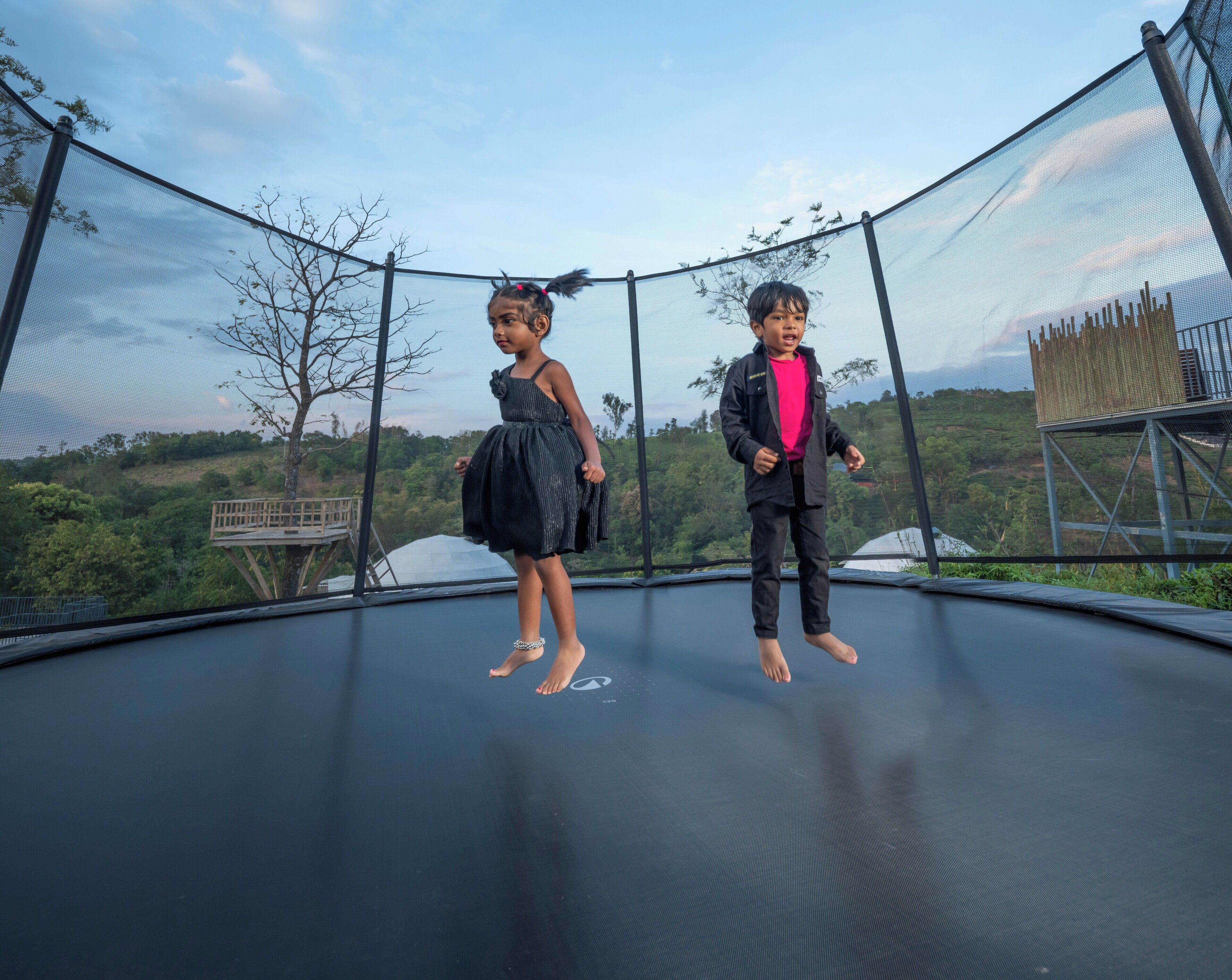 Two children standing on a trampoline with a scenic view of trees and hills in the background.