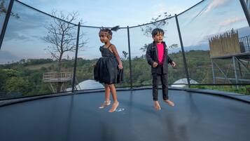 Two children standing on a trampoline with a scenic view of trees and hills in the background.