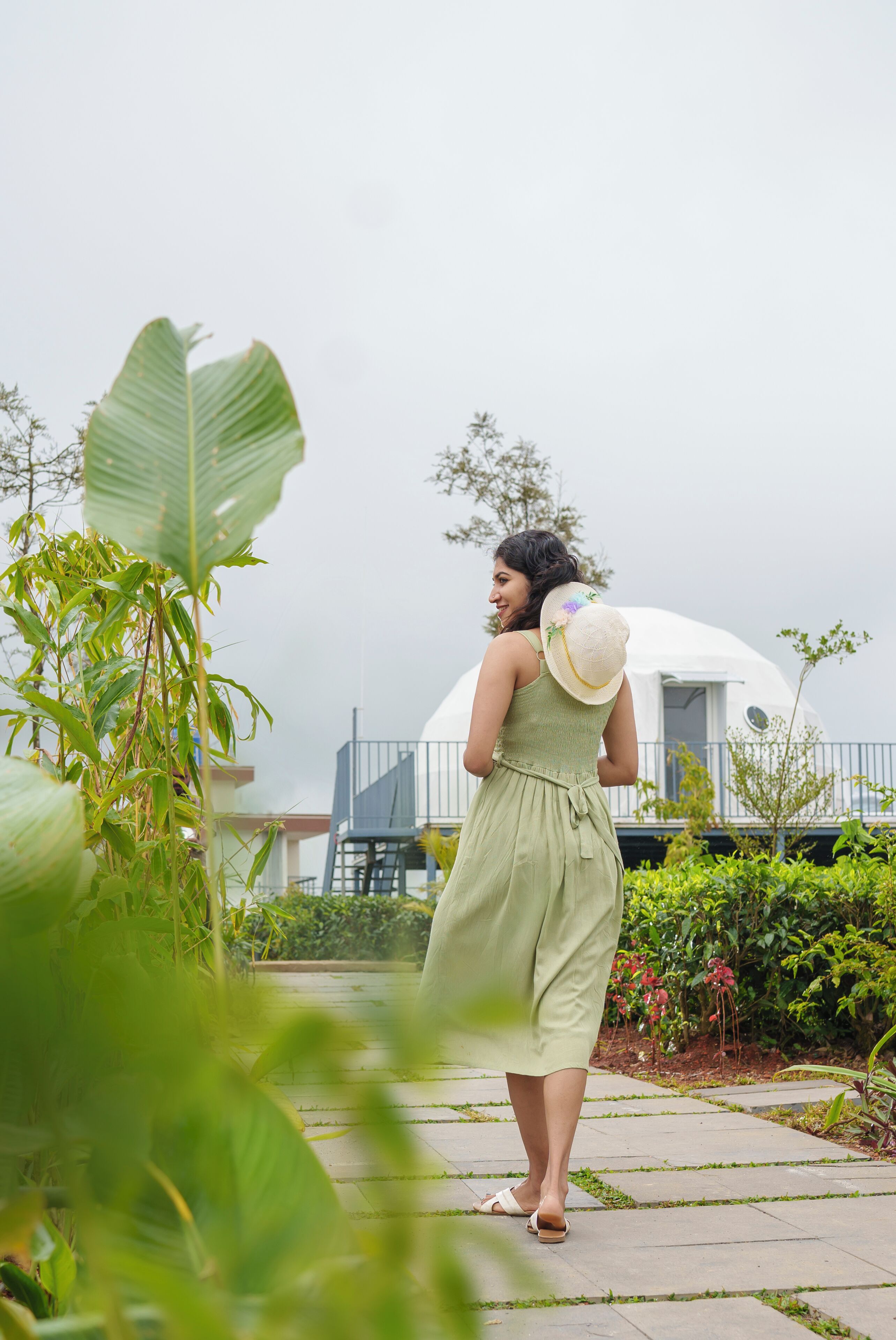 A woman in a green dress walking on a pathway with lush greenery and a white dome-shaped building in the background.