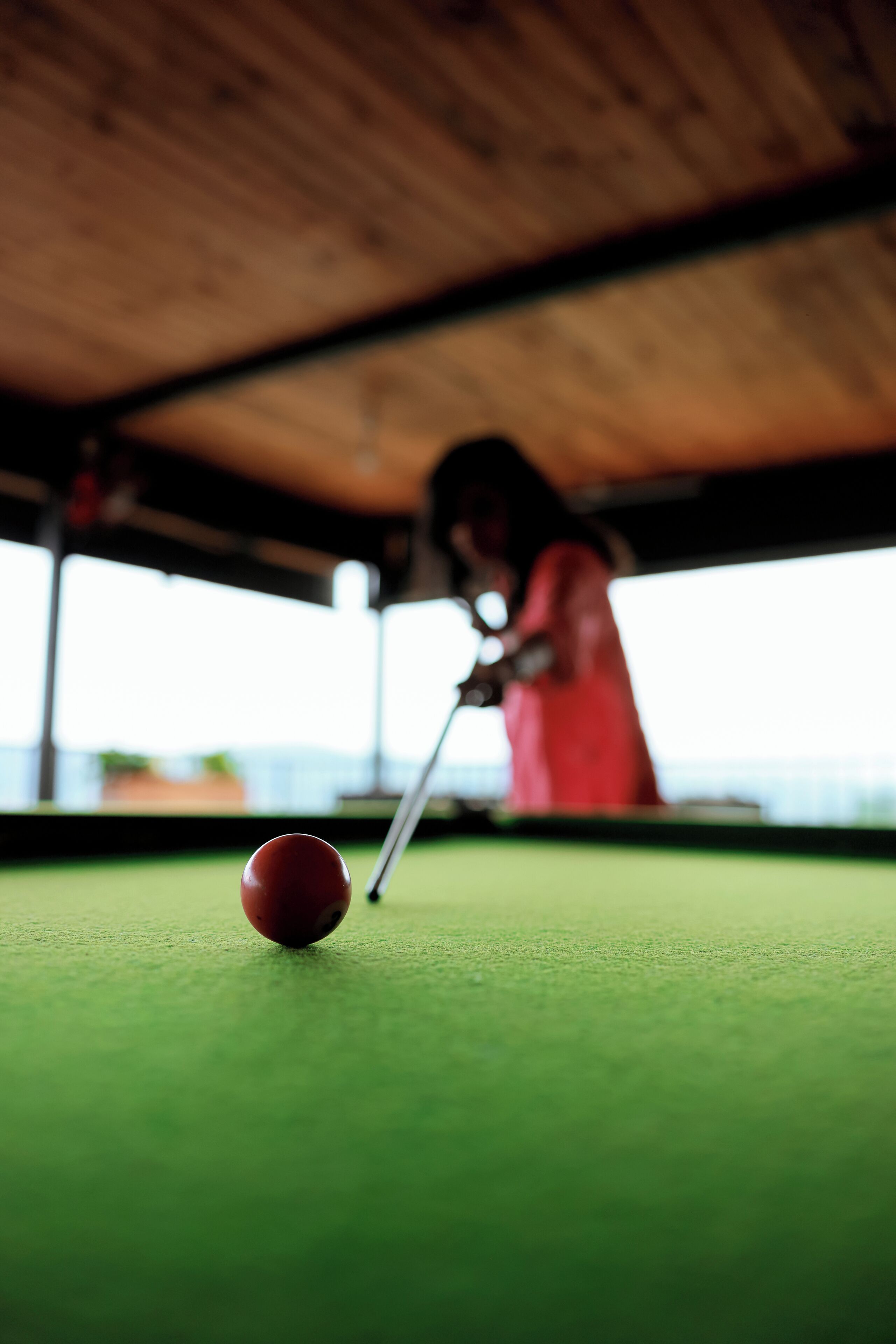 A billiards table with a red ball in focus, a cue stick, and a person in the background.