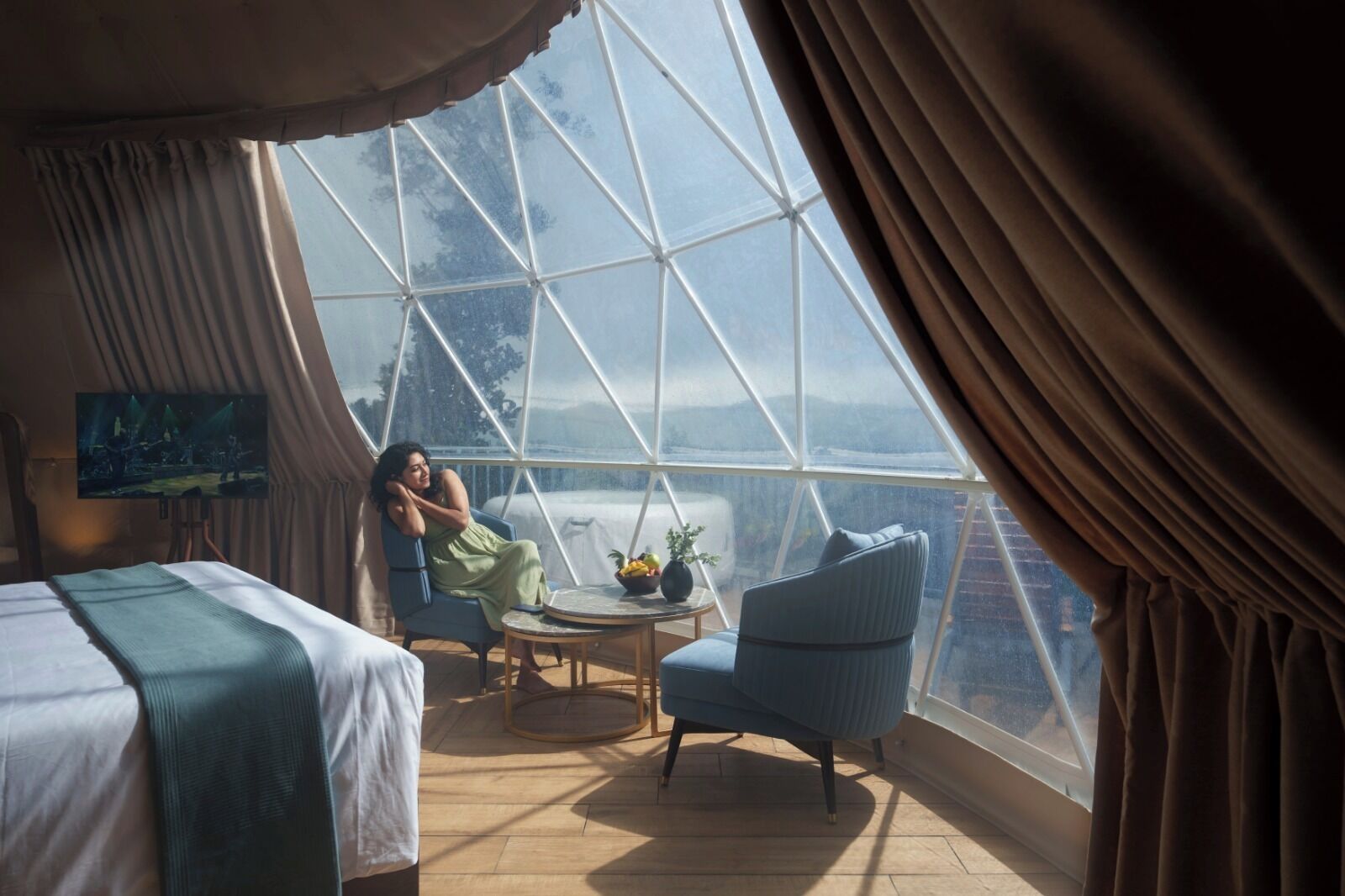 A room with a large window, a bed, a chair, a small table with a plant, and a painting on the wall.
