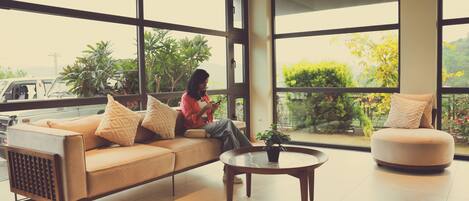 A modern living room with a sofa and armchair, a coffee table, and large windows offering a view of greenery and a vehicle outside.