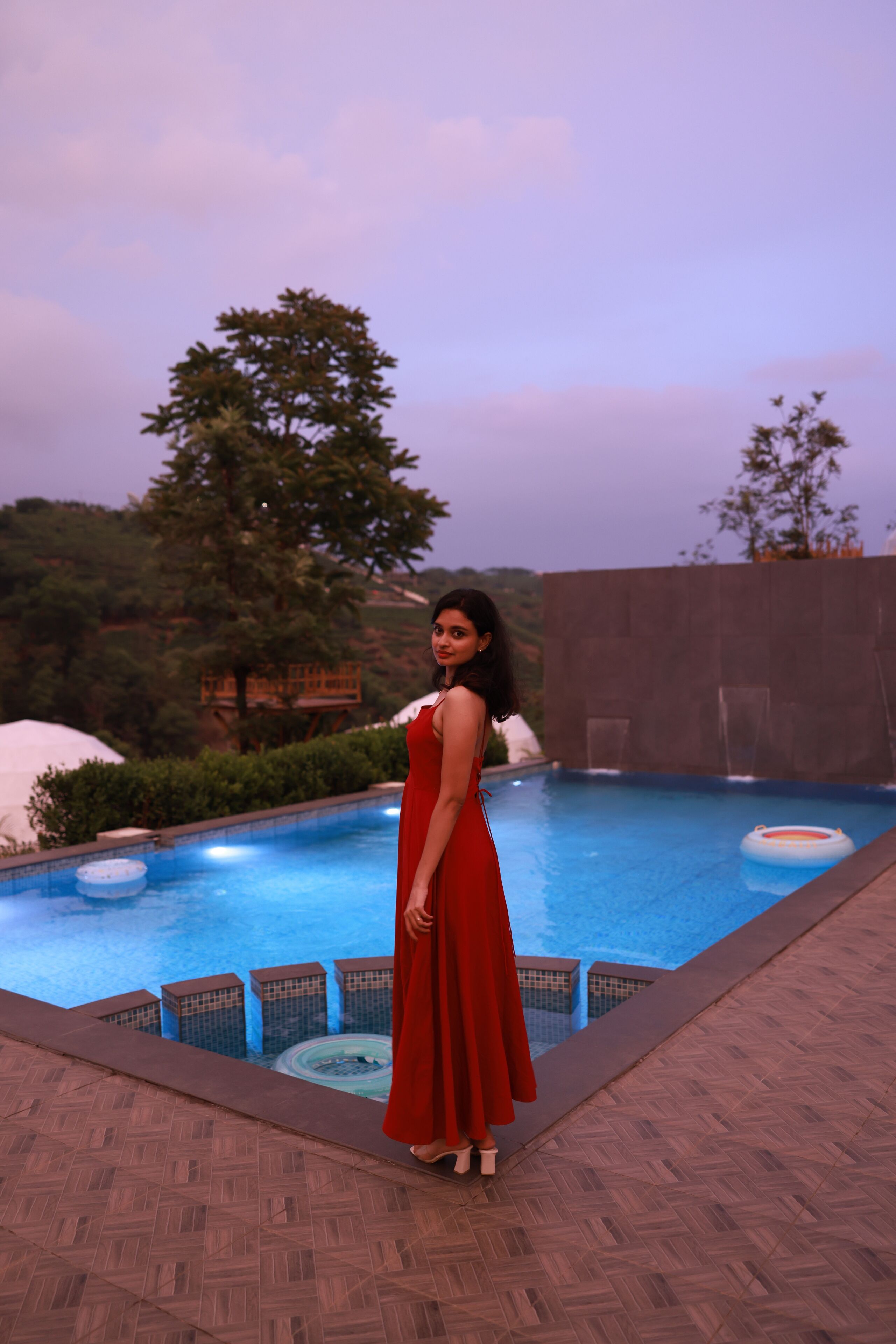 A woman in a red dress standing by a pool with a view of trees and a stone wall.