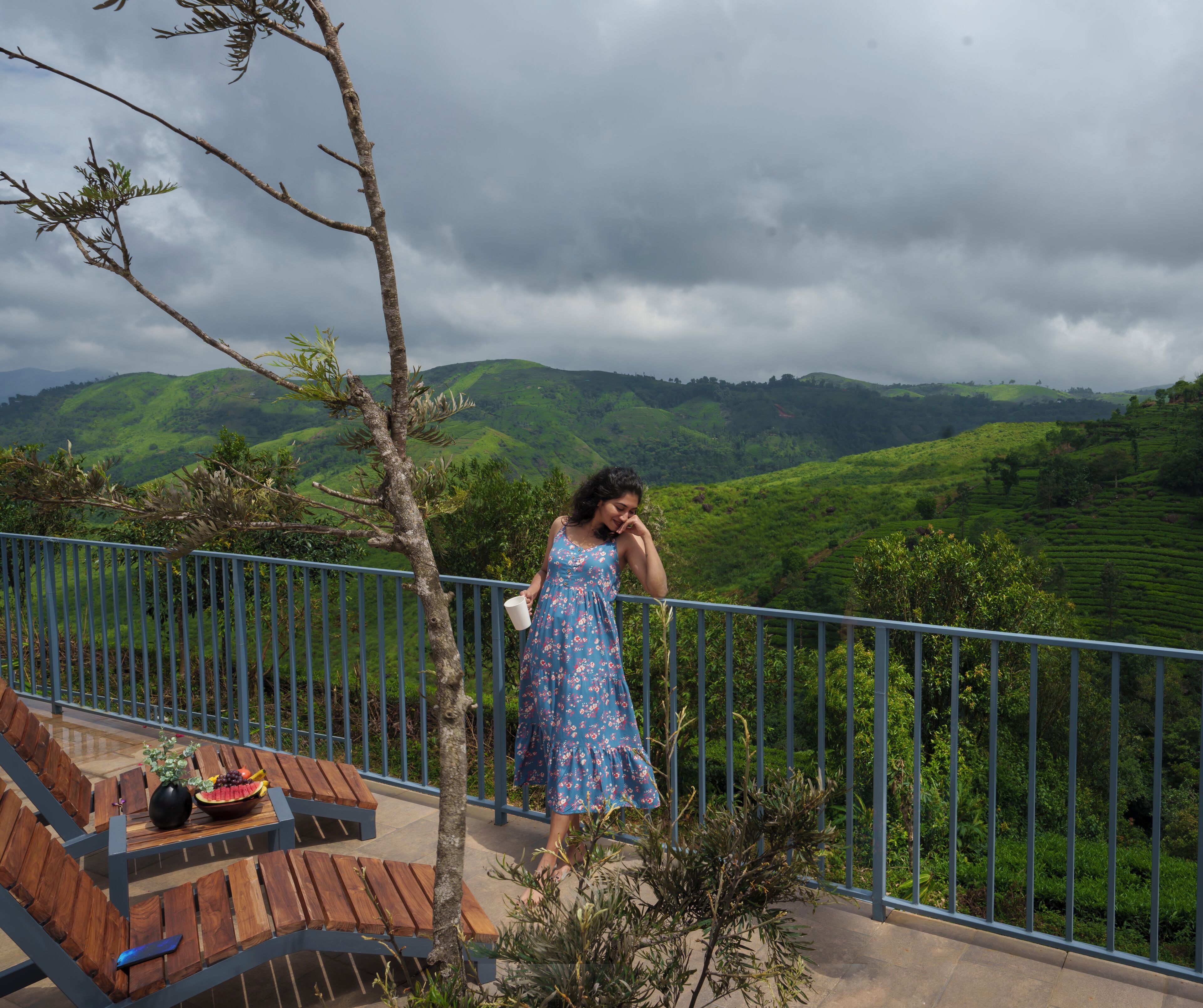 Une femme vêtue d’une robe bleue se tient sur un balcon, d’où l’on peut admirer des collines verdoyantes.