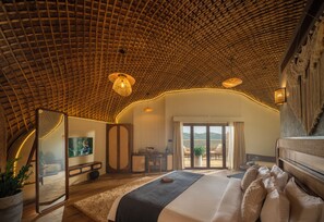 A spacious bedroom with a curved ceiling, a large bed, a television, and a balcony with outdoor seating. - Monticle Glamp (Peermade)