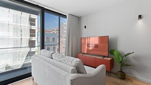 A modern living room with a grey sofa, a flat-screen TV on a wooden cabinet, and a large window overlooking a cityscape. - GuestReady - Age of Discoveries - Gama (Porto)