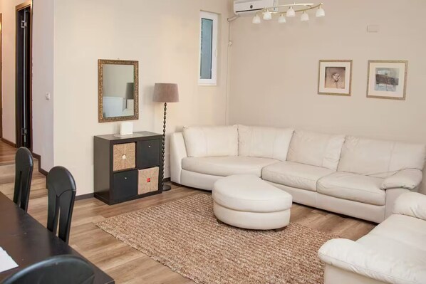 A living room with a white sofa, a small ottoman, a dark wooden side table with a lamp, and a large mirror.