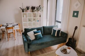 Un salón con un sofá verde oscuro, una mesa de comedor con sillas, una estantería blanca con plantas y una guitarra.