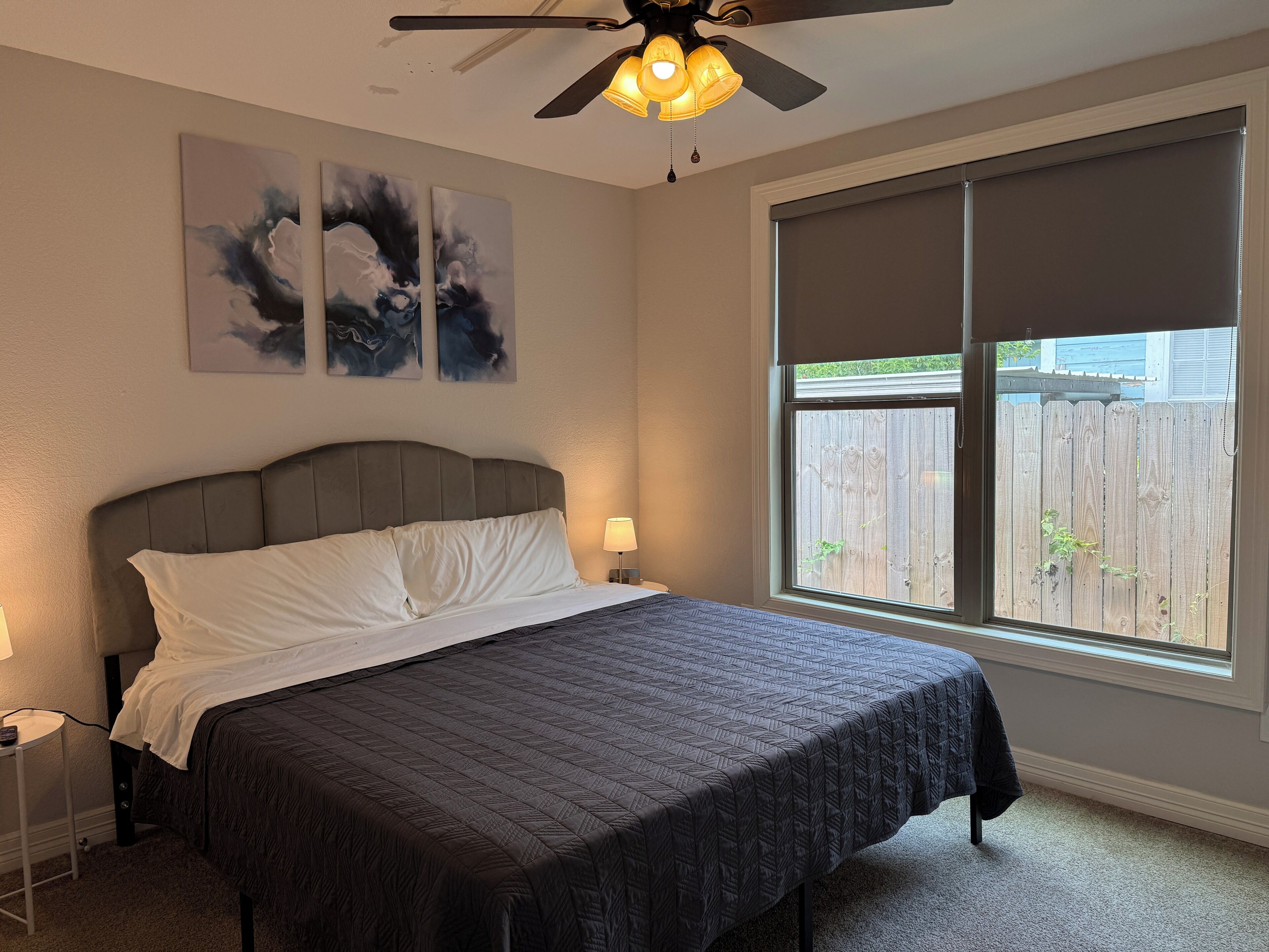 A bedroom with a bed, a ceiling fan, artwork on the wall, and a window with a view of a wooden fence.