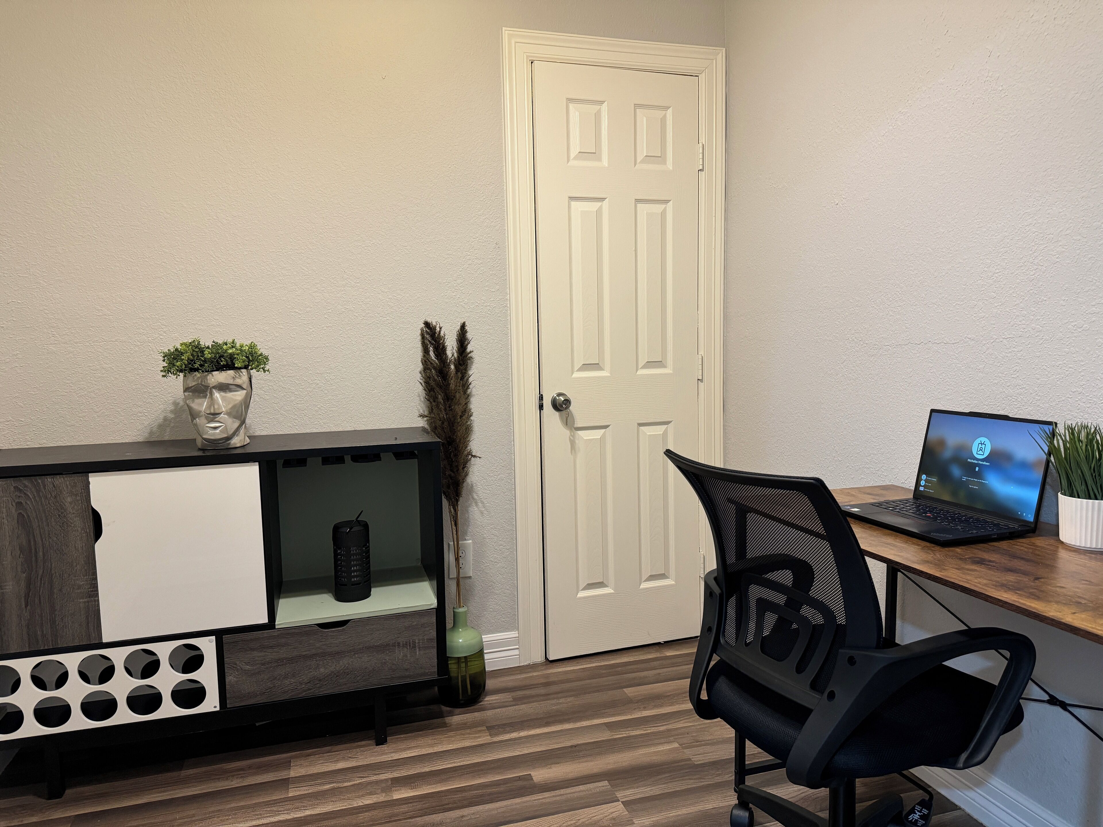 A room with a wooden desk, a laptop, a chair, a decorative vase, and a door.