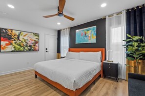 Een moderne slaapkamer met een houten vloer, een bed met wit beddengoed, een plafondventilator en twee schilderijen aan de muur.