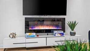 Ein modernes Wohnzimmer mit flachbildfernseher an der Wand, einem Kamin aus Glas, einer weißen Unterhaltungsecke mit Schubladen und einer Topfpflanze.