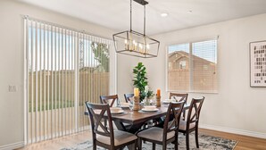A dining area with a wooden table, six chairs, a chandelier, and a large window with blinds.